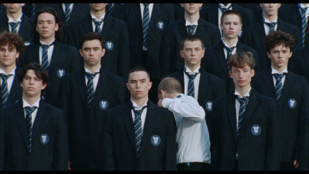 Group of young men in dark blazers and striped ties standing in a formal lineup with blue badges on their jackets.