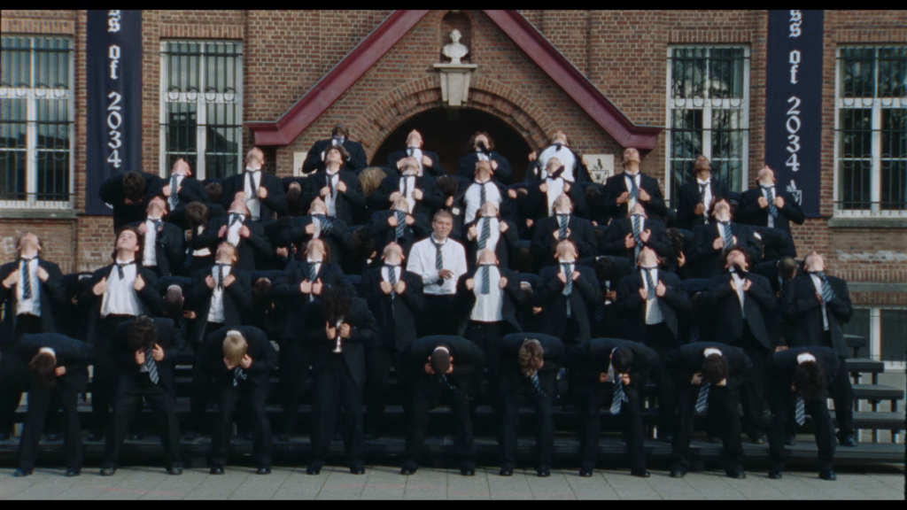 Large group of men in dark suits and striped ties on outdoor steps in front of a brick campus building, many tilting their heads back and bowing their bodies as if singing or cheering.