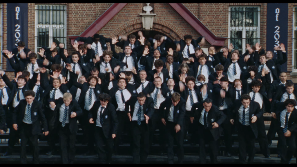 Large group of students in navy blazers and striped ties raising hands on stairs in front of a brick school building.