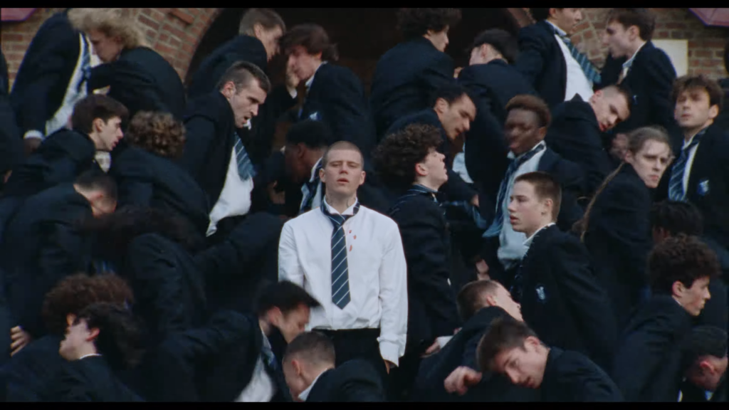 Large crowd of young men in dark suits and striped ties crowding together near a brick entrance, with a central figure in a white shirt and loosened tie standing at the front and shoulder-bloodstain visible on his chest.