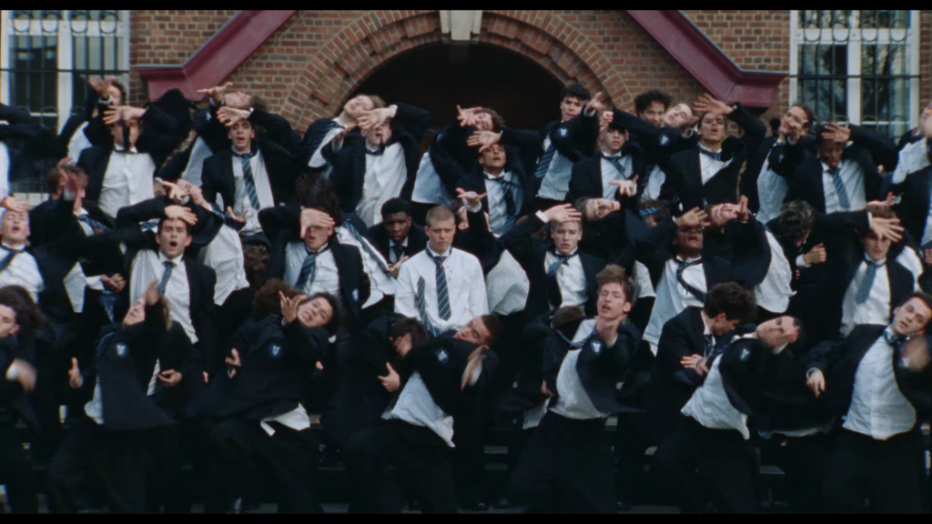 Large group of students in uniforms performing a synchronized dance with hands over their eyes in front of a brick building.