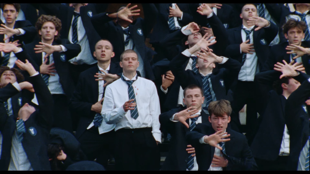 Group of students in navy blazers and striped ties raising their arms in a synchronized gesture at a crowded event