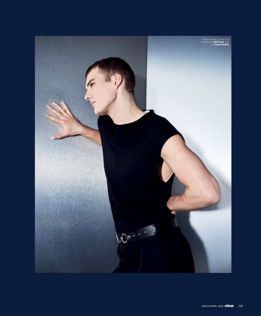 Male model in a black sleeveless shirt and belt leans against a metallic wall, one hand pressed on the surface.