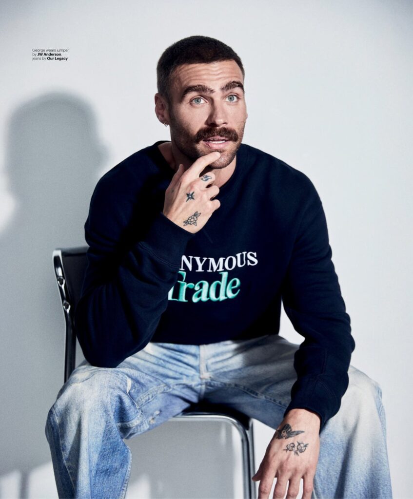 Man with short dark hair and tattoos sits on a metal chair, touching his chin, wearing a navy sweatshirt with turquoise 'Trade' text.