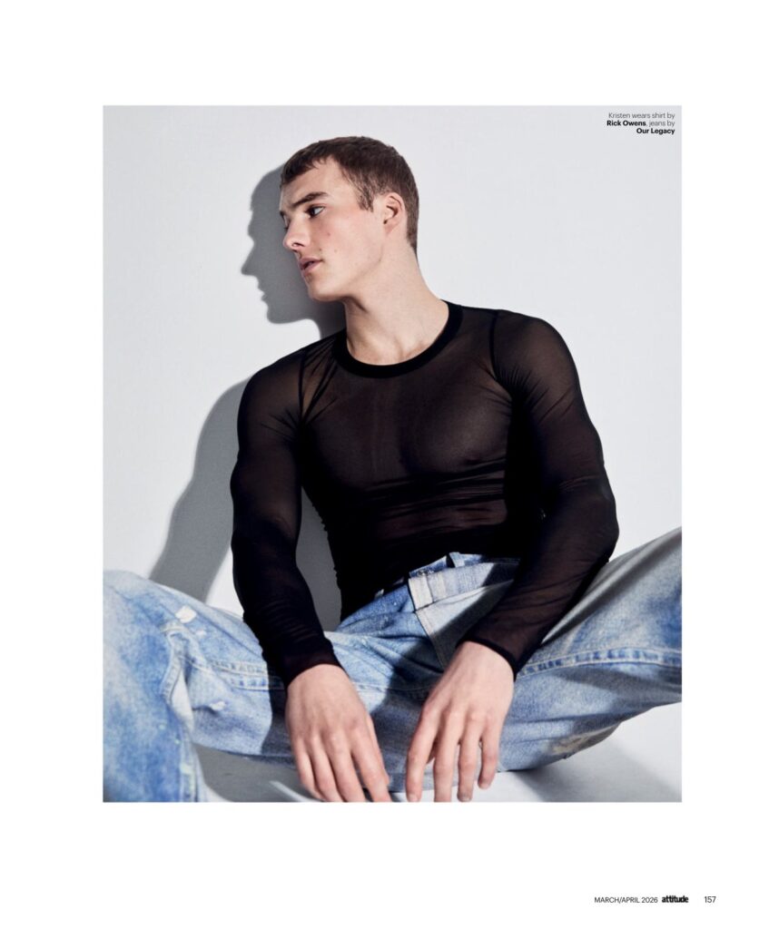 Male model wearing a sheer black long-sleeve top and light-wash jeans, seated against a light background in a fashion shoot to showcase the outfit.
