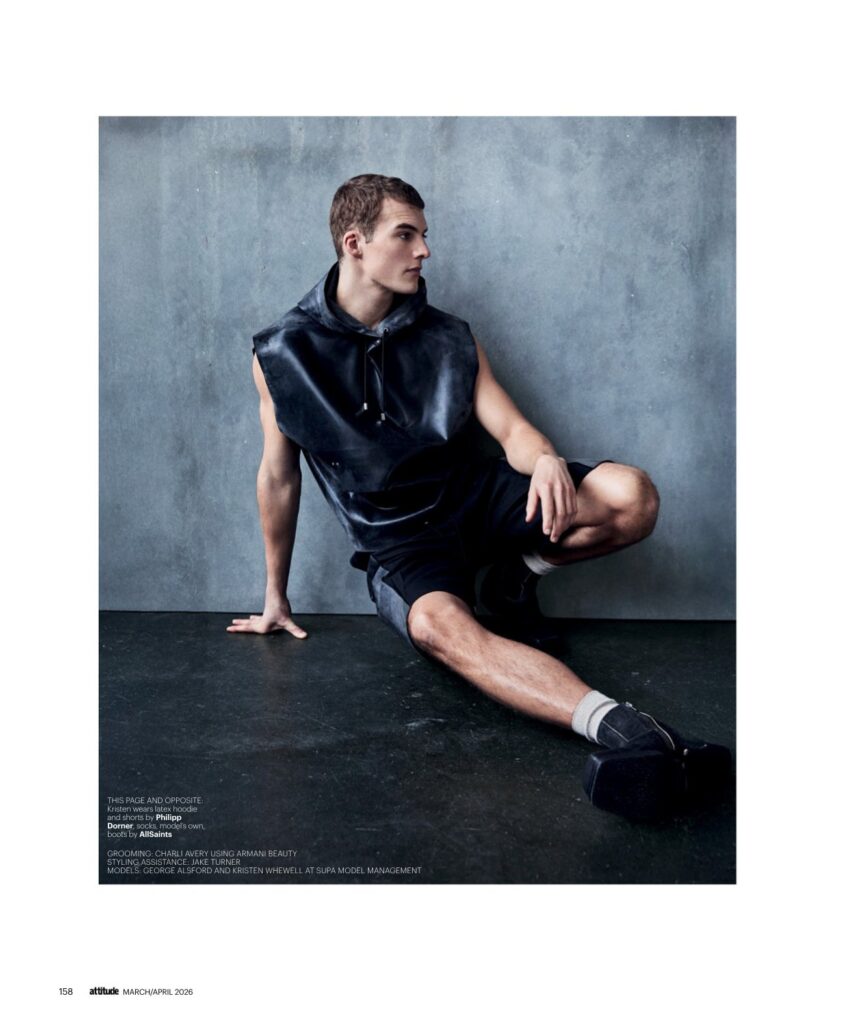 Male model wearing a sleeveless black latex hoodie and shorts sits on a dark studio floor against a gray textured wall, posing for a fashion shoot.
