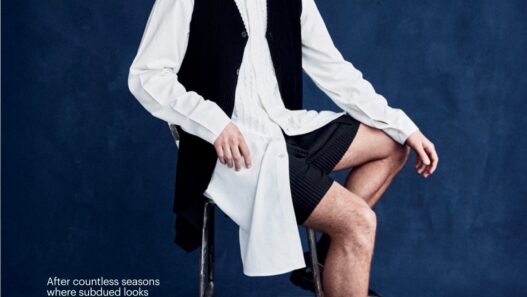 Male fashion model seated on a metal chair against a dark blue studio backdrop, wearing a white shirt with a black vest and shorts; editorial page with text blocks along left and small captions on right.