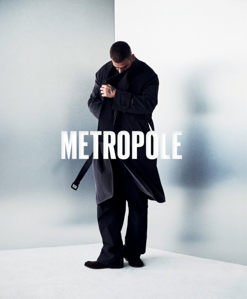 Man in a dark coat adjusting his collar in front of a pale, frosted backdrop with the word METROPOLE across the image (poster-style promotional photo).