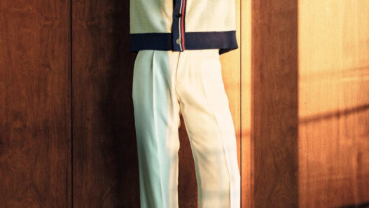 Man in a cream cardigan with navy trim and light mint trousers poses with arms behind head against a wooden wall, wearing mesh shoes.