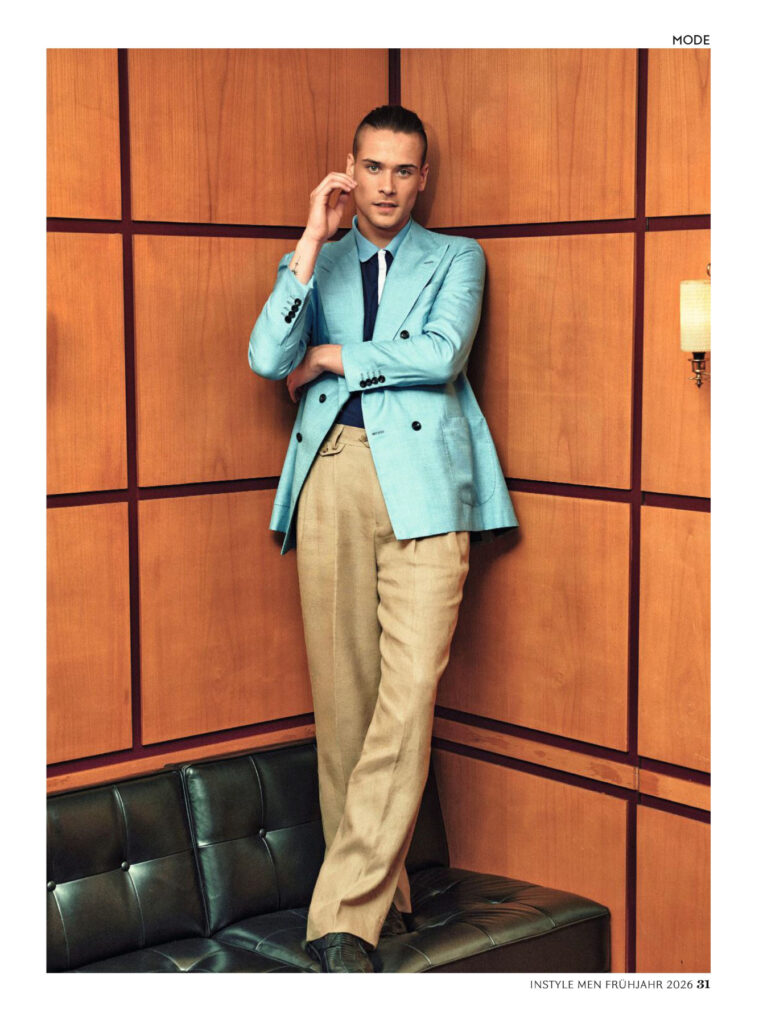 Male model in a turquoise blazer and beige pants posing against a wood-paneled wall for a fashion shoot.