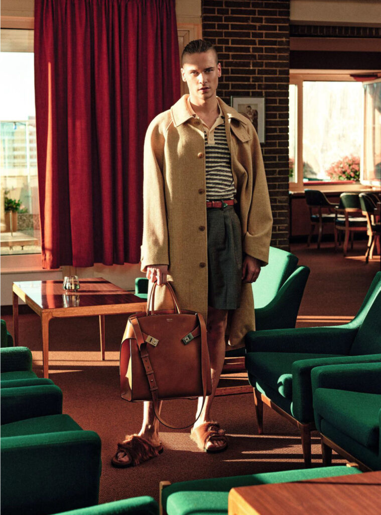 Man standing in a retro hotel lounge, wearing a tan overcoat, striped shirt, gray shorts, and carrying a brown leather bag.