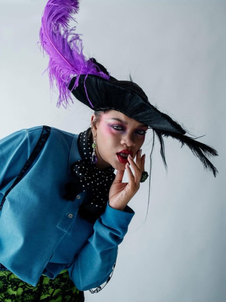 Fashion portrait of a person in a dramatic black hat with a purple feather, bold pink makeup, and a blue shirt.