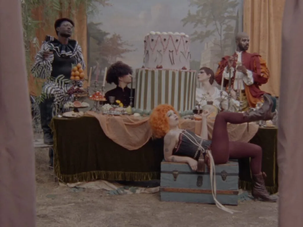 People in colorful period costumes around a feast and a tall decorated cake on a table outdoors, as if at a circus or masquerade party.
