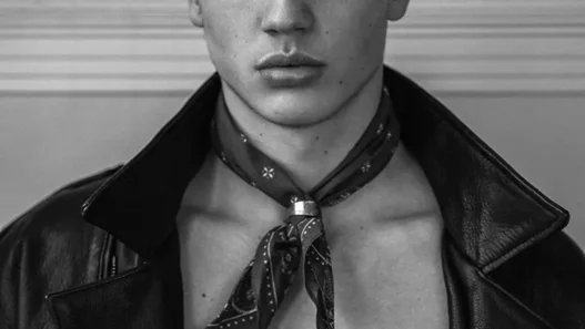 Black-and-white portrait of a male model wearing a leather jacket, a cap, and a neck scarf, looking directly at the camera.