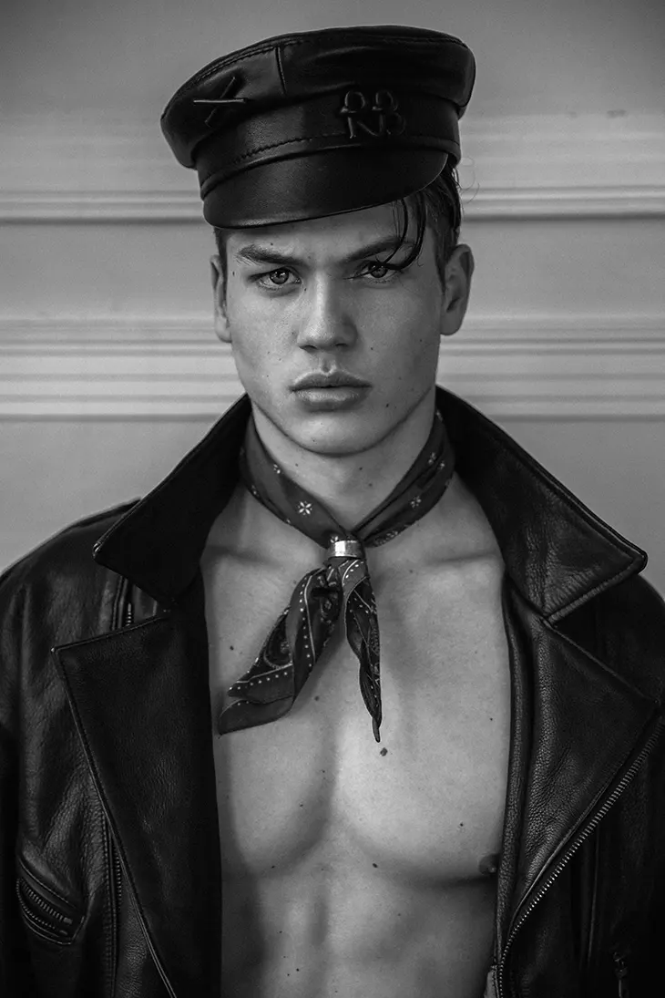 Black-and-white portrait of a male model wearing a leather jacket, a cap, and a neck scarf, looking directly at the camera.