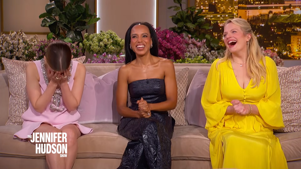 Three women sit on a beige couch on a talk-show set; the left woman covers her face in a pink dress, the middle woman smiles in a black strapless dress, and the right woman laughs in a bright yellow dress.