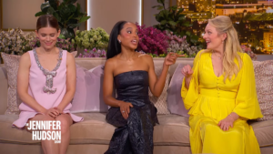 Three women sit on a beige couch on a talk-show set with bright floral arrangements in the background, part of The Jennifer Hudson Show logo visible in the corner.