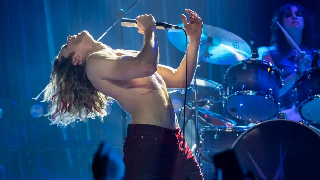 Shirtless male lead singer grips a microphone, head tilted back mid‑song on a blue-lit stage; drummer behind him.