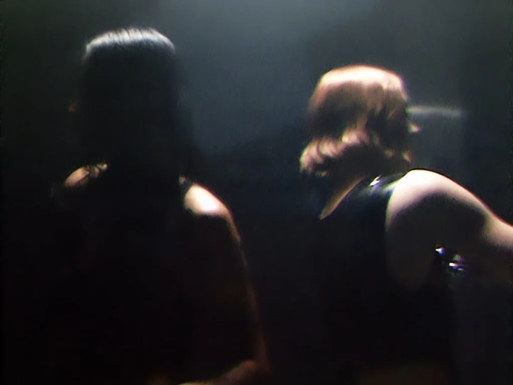 Dark, blurry concert scene with several indistinct silhouettes and raised arms in a dim venue.