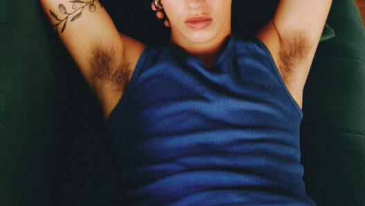 Relaxed young man lying on a green sofa, wearing a blue sleeveless shirt, arms behind his head and visible arm tattoos.