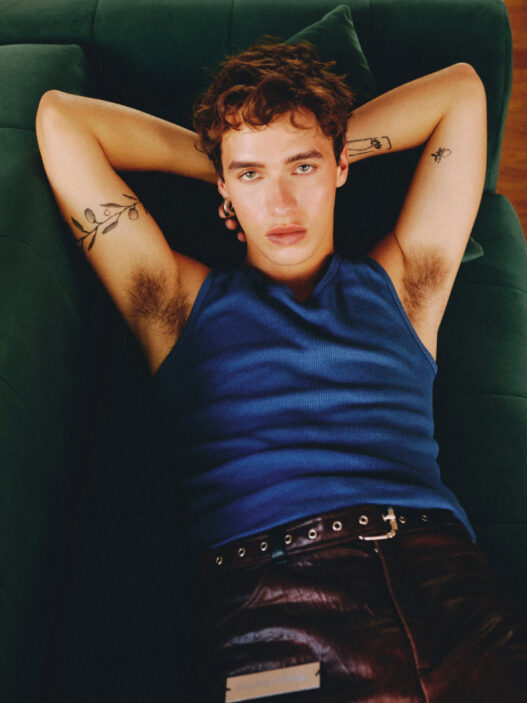 Relaxed young man lying on a green sofa, wearing a blue sleeveless shirt, arms behind his head and visible arm tattoos.