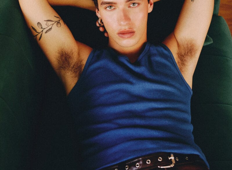 Relaxed young man lying on a green sofa, wearing a blue sleeveless shirt, arms behind his head and visible arm tattoos.