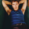 Relaxed young man lying on a green sofa, wearing a blue sleeveless shirt, arms behind his head and visible arm tattoos.