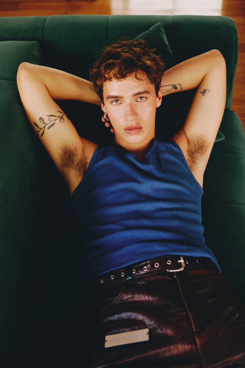 Relaxed young man lying on a green sofa, wearing a blue sleeveless shirt, arms behind his head and visible arm tattoos.