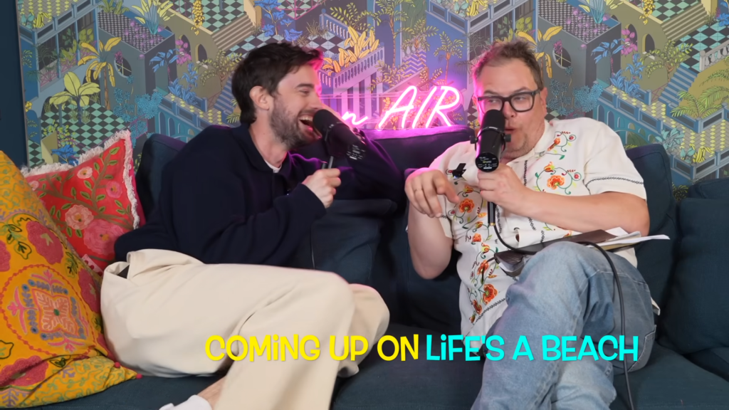 Two men sit on a blue couch recording a podcast, speaking into handheld microphones with a neon 'ON AIR' sign behind them on a colorful patterned wall.