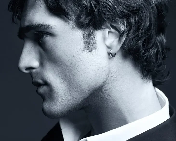 Side profile of a young man in a suit, wearing a hoop earring, in a moody black-and-white portrait.