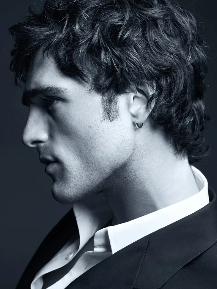 Side profile of a young man in a suit, wearing a hoop earring, in a moody black-and-white portrait.