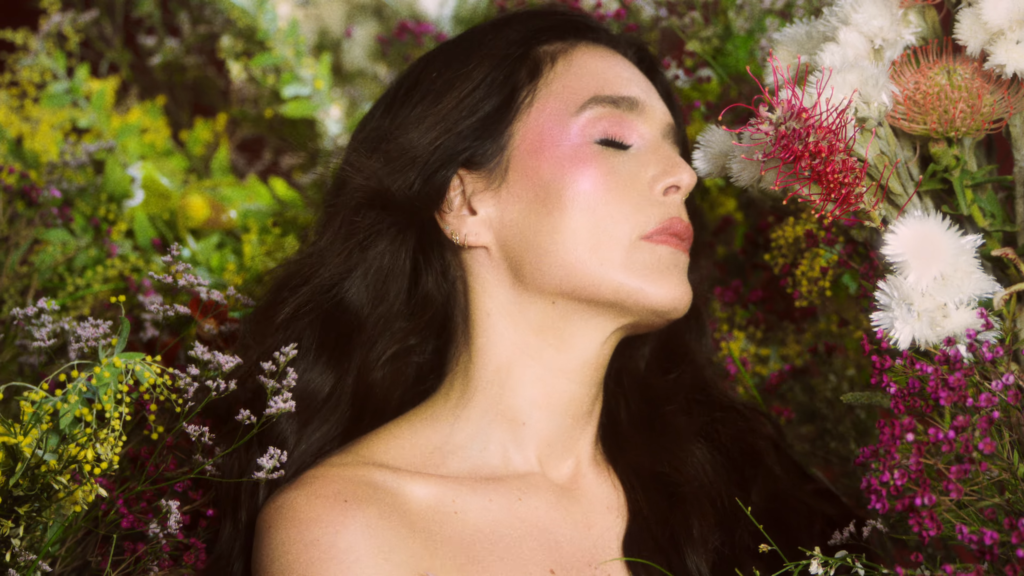 Portrait of a woman with long dark hair amid colorful flowers, eyes closed, head tilted back as if savoring the scent.