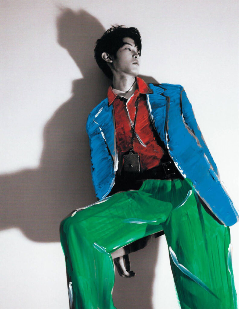 Stylized fashion portrait: person in a bold blue jacket, red shirt, and bright green pants against a plain background, looking to the side.