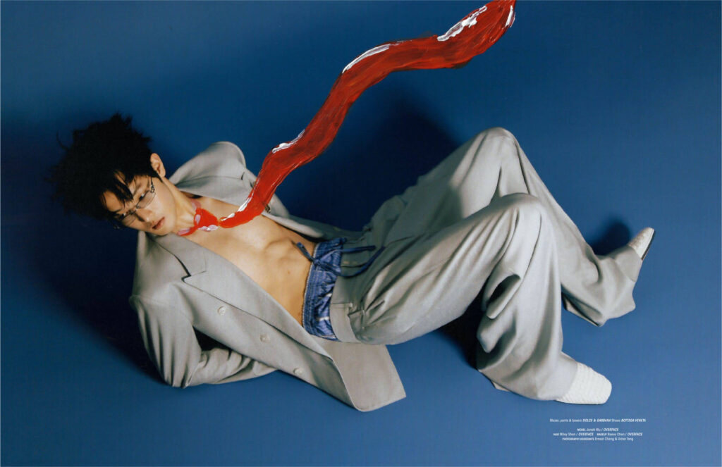Young man in a gray suit lying diagonally on a blue backdrop, with a red painted ribbon draped across his chest.