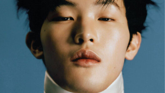 Close-up portrait of a person with tousled black hair, wearing a white bow tie and high collar against a blue background.