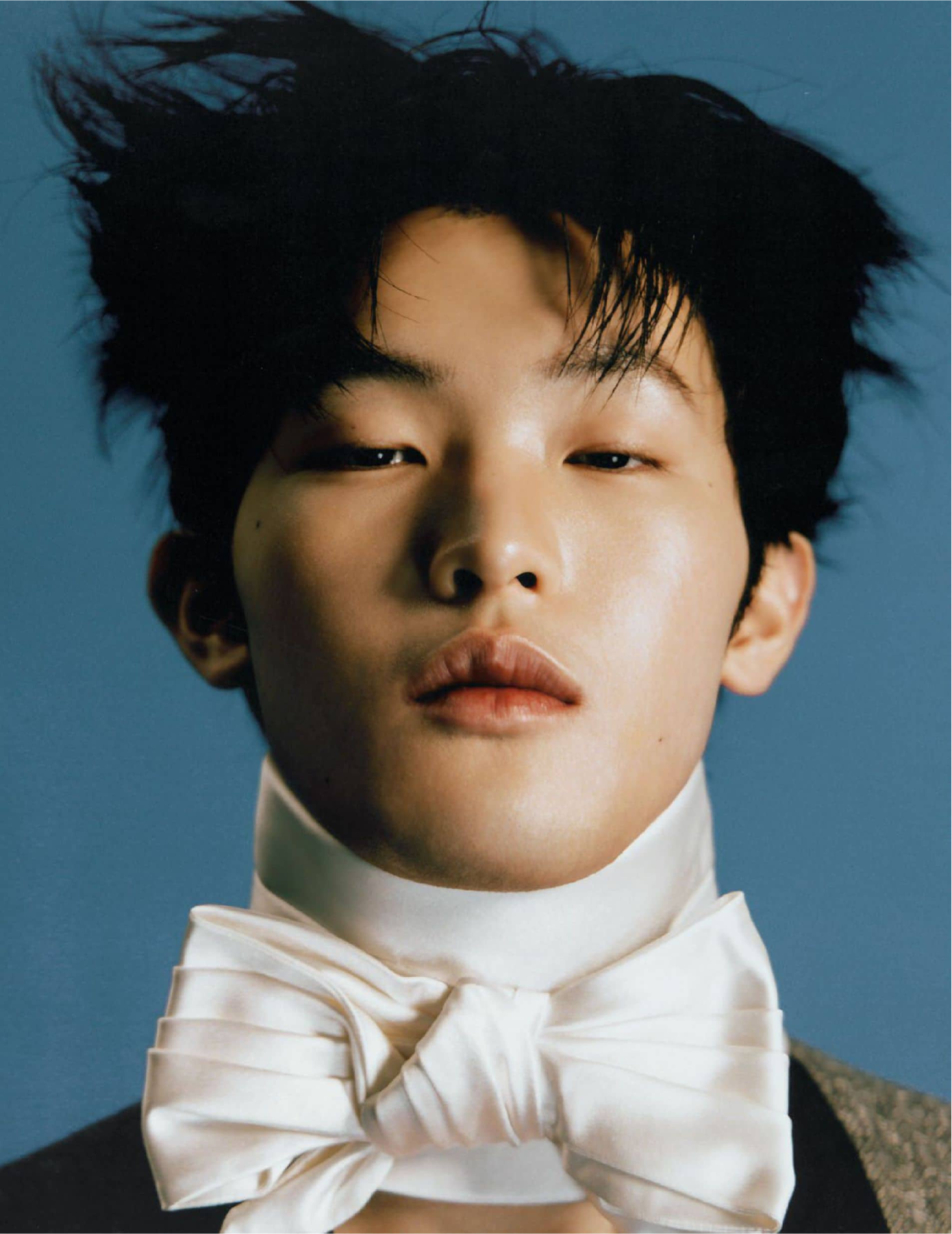 Close-up portrait of a person with tousled black hair, wearing a white bow tie and high collar against a blue background.