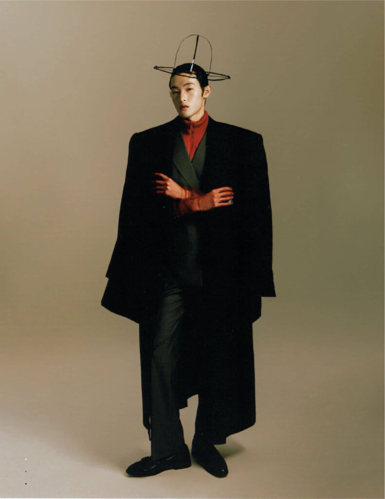 Model wearing a long black coat over a red turtleneck, with a circular wire headpiece, posed against a beige studio background.| height=auto