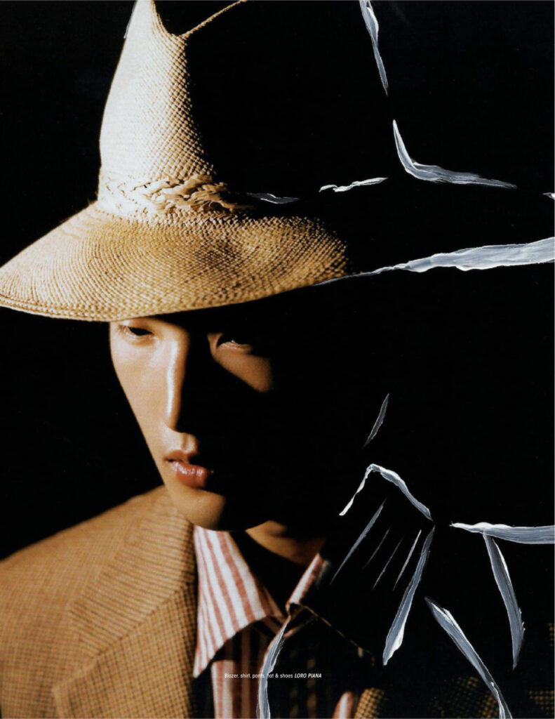 Close-up of a person wearing a large straw hat and tan blazer against a black background, with torn white paper edges across the image.