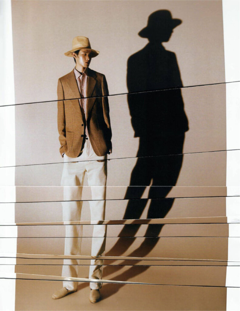 Man in a tan blazer, striped shirt, white pants, and a hat stands with hands in pockets, with a large shadow on the wall beside him.