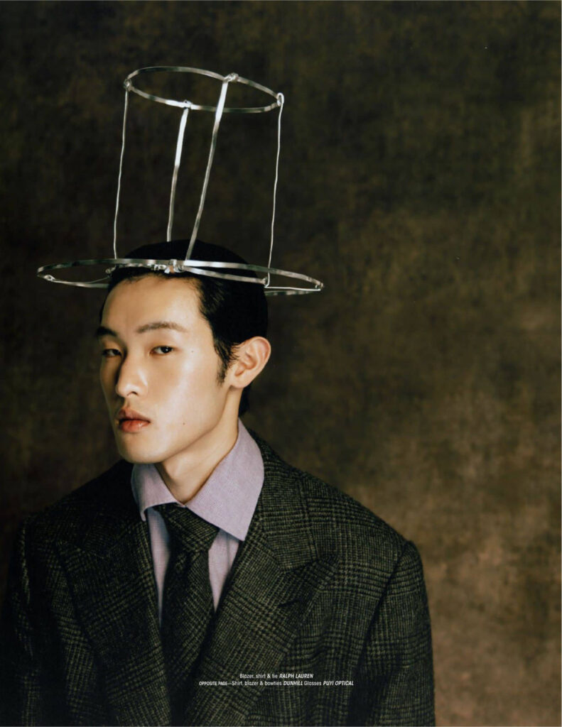 Portrait of a man in a tweed blazer and lavender shirt wearing a sculptural hat made of metal hoops against a dark backdrop.