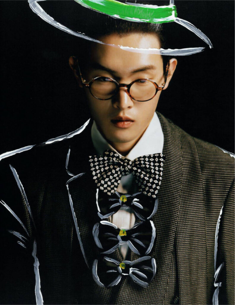 Fashionably dressed man in a brown check blazer, white shirt, and geometric bow tie, wearing round glasses against a dark background with white painted streaks.
