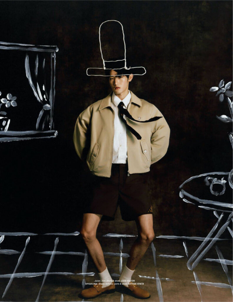 Male model in a beige jacket, white shirt, dark tie, and brown shorts standing with hands in pockets, chalk-drawn hat outline around his head in a dark studio.