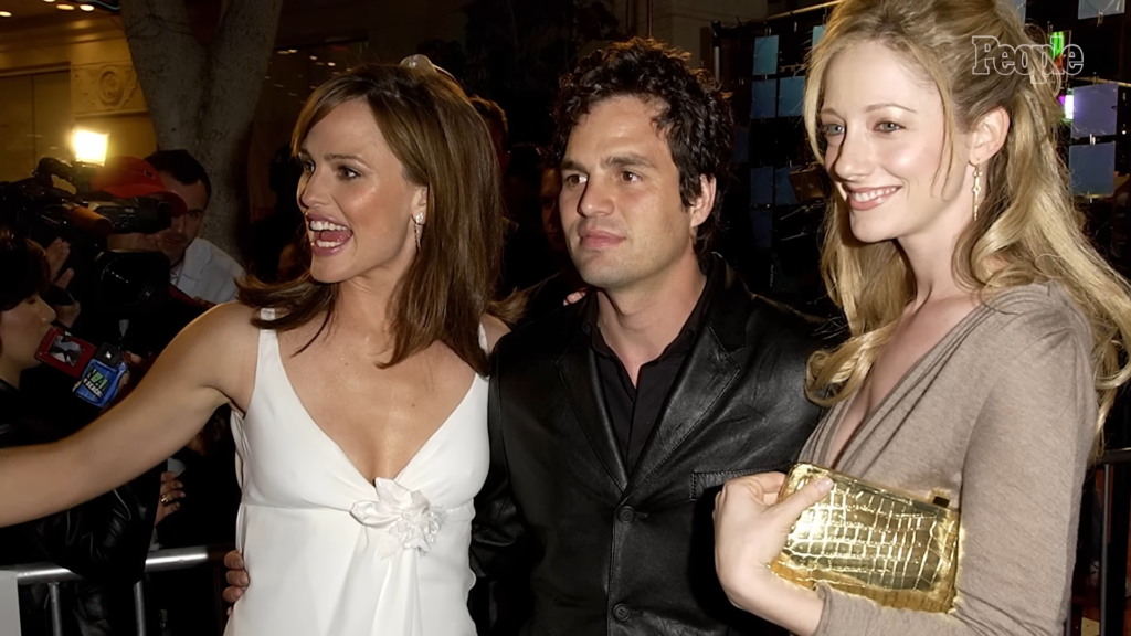 Three people pose for cameras at a red-carpet event: a woman in a white dress on the left, a man in a black suit center, and a woman in beige with a gold clutch on the right.