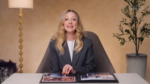 Woman in a gray blazer sits at a white table with an open photo album, speaking to the camera in a warm studio setting.