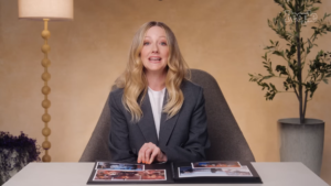 Woman in a gray blazer sits at a white table with an open photo album, speaking to the camera in a warm studio setting.