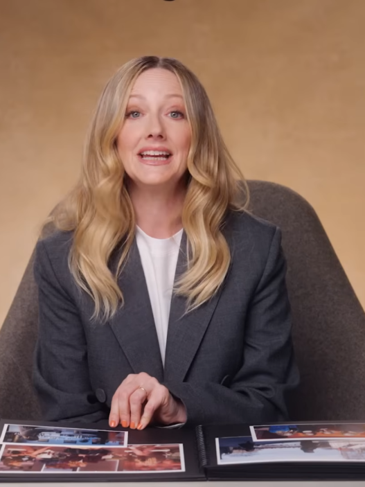 Woman in a gray blazer sits at a white table with an open photo album, speaking to the camera in a warm studio setting.
