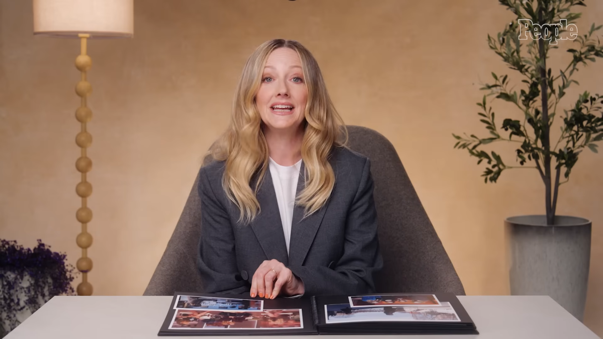 Woman in a gray blazer sits at a white table with an open photo album, speaking to the camera in a warm studio setting.