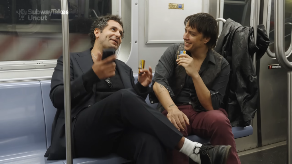 Two men sit in a subway car, smiling and chatting; one in a dark suit holding a phone, the other in a dark shirt and maroon pants holding a snack bar