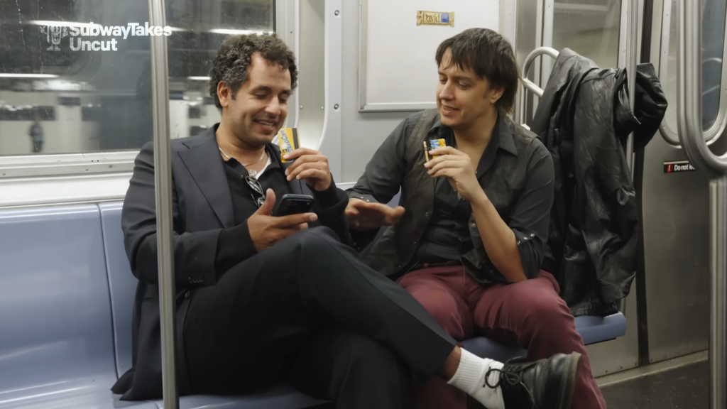 Two men seated on a subway train, sharing candy bars and chatting.