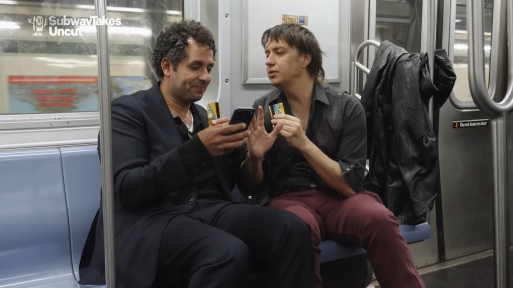 Two men sit closely on a subway car, sharing snacks and looking at a smartphone together.